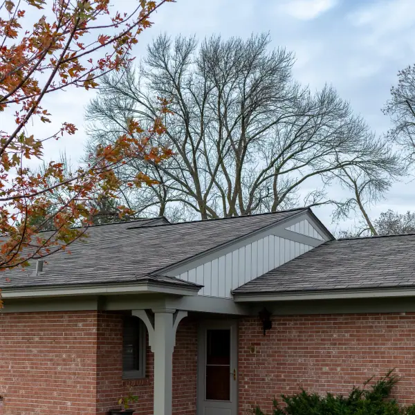 Covered brick entryway with a gray storm door beneath a newly shingled roof and board-and-batten gable.