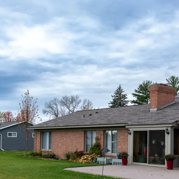 Rear elevation of a brick ranch home with new shingles, a brick chimney and a sliding patio door onto a lawn.