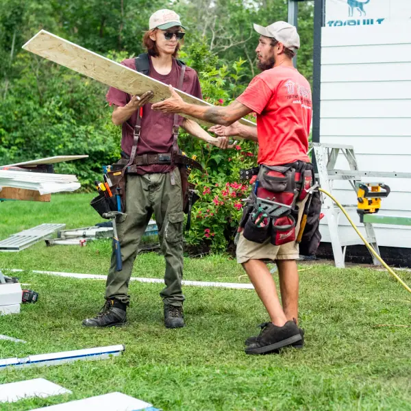 One crew member hands a length of sheathing board to another beside a partially sided wall.