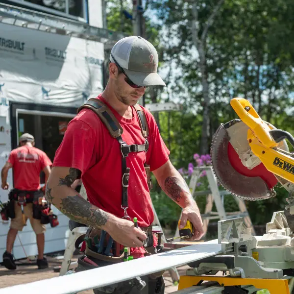 Installer measuring white trim at a miter saw station, with the house wrapped in weather barrier behind.
