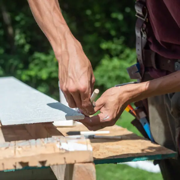 Close-up of a worker's hands scoring a white trim board laid across wooden sawhorses.